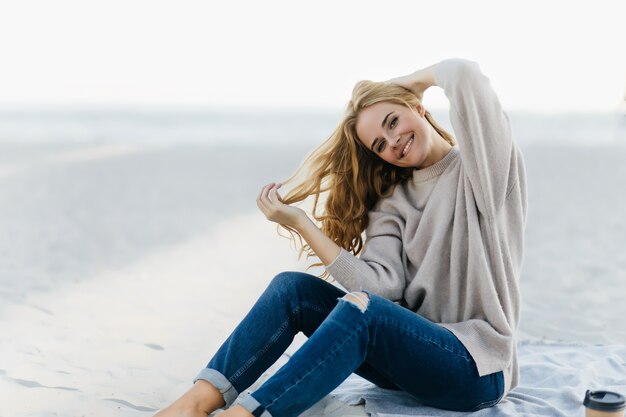 Donna allegra che posa emotiinal alla spiaggia di autunno. Ritratto all'aperto di donna abbastanza riccia in jeans che si siedono nella sabbia.
