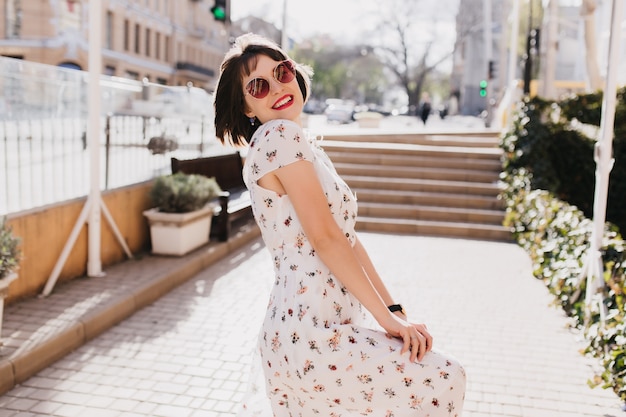 Donna allegra che ride in occhiali da sole alla moda divertendosi nella soleggiata giornata estiva. Outdoor ritratto di piacevole ragazza con taglio di capelli corto indossa un abito bianco nella calda mattina di primavera.