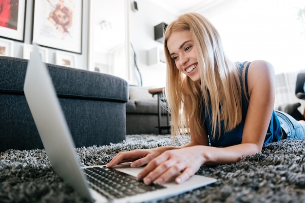 Donna allegra che si trova sul tappeto e che per mezzo del computer portatile a casa