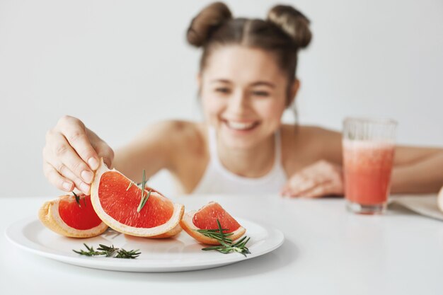 Donna allegra che sorride prendendo fetta di pompelmo dal piatto che si siede alla tavola sopra la parete bianca. Concetto di fitness sano. Focus sul cibo.