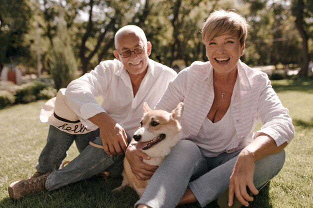 Donna allegra con l'acconciatura alla moda bionda in camicia rosa e jeans ridendo, abbracciando corgi e seduto con un uomo dai capelli grigi in occhiali nel parco.