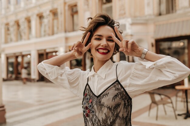 Donna allegra con labbra rosse che mostra segni di pace in città. Donna moderna con i capelli corti in camicetta bianca con pizzo nero che sorride alla strada.