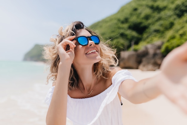 Donna allegra con pelle abbronzata che fa selfie all'isola tropicale. Foto all'aperto di giovane donna estatica in occhiali da sole alla moda di scattare una foto di se stessa alla spiaggia sabbiosa.