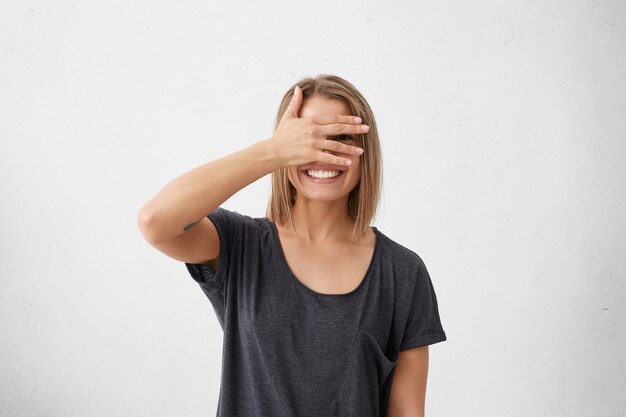 Donna allegra con uno sguardo timido che sbircia tra le dita. Giovane femmina carina imbarazzata con pettinatura bob che copre il viso con la mano che guarda in modo confuso sorridente ampiamente