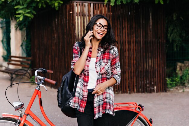 Donna allegra con zaino in pelle nera, parlando al telefono per strada. Graziosa ragazza dai capelli neri in piedi accanto alla bicicletta rossa.