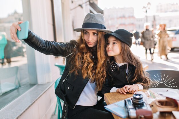 Donna allegra dai capelli castani elegante in cappello di feltro che fa selfie con la figlia affascinante in attesa del caffè nella caffetteria.