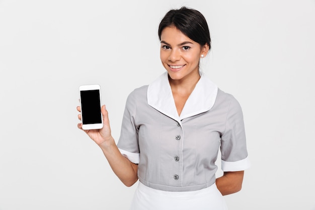 Donna allegra del brunette in uniforme grigia che mostra schermo mobile in bianco mentre levandosi in piedi