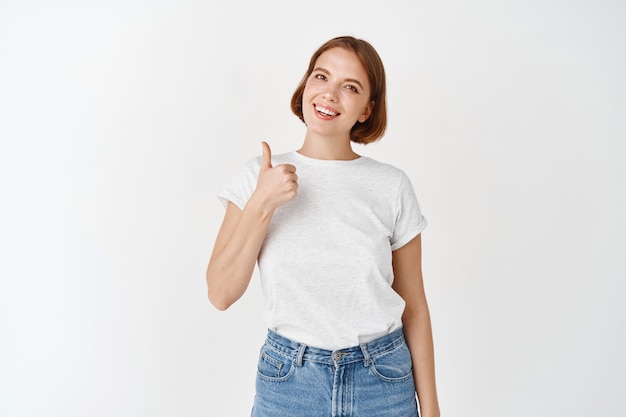 Donna allegra in maglietta che mostra pollice in su e sorride, approva e piace. La ragazza con i capelli corti e l'aspetto naturale loda il buon lavoro, in piedi sul muro bianco white