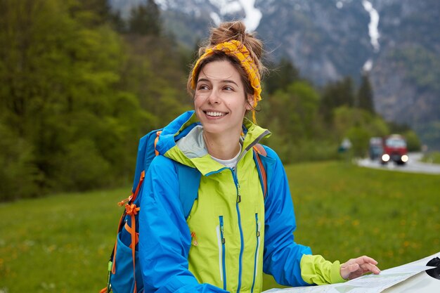 Donna allegra sognante vestita in abbigliamento sportivo, utilizza la mappa turistica per trovare il percorso giusto, indossa lo zaino