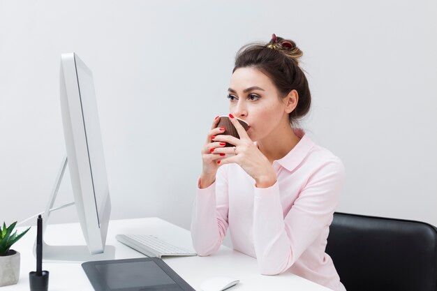 Donna allo scrittorio che beve caffè e che esamina computer