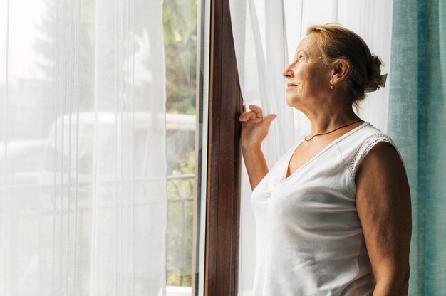 Donna anziana a casa durante la pandemia guardando attraverso la finestra