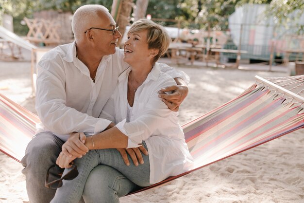 Donna anziana allegra con l'acconciatura fresca bionda in camicetta e jeans che si siede sull'amaca a quadri e abbraccia con sorridente uomo dai capelli grigi sulla spiaggia