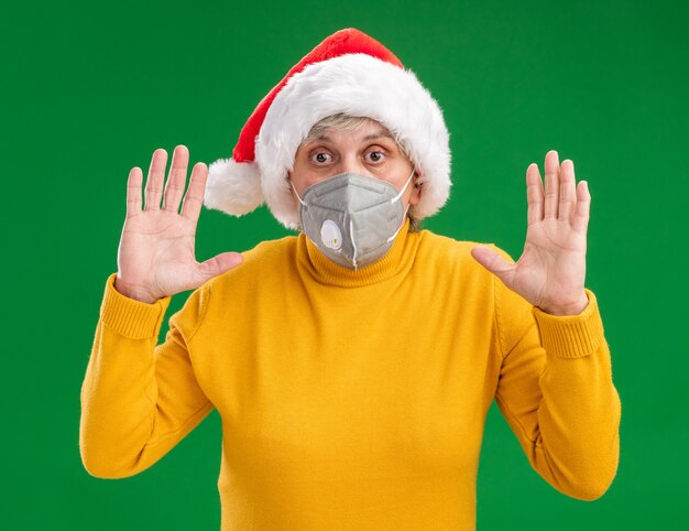 Donna anziana ansiosa con cappello da Babbo Natale che indossa una maschera medica in piedi con le mani alzate isolate sul muro verde con spazio copia