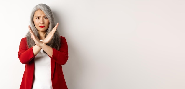 Donna anziana asiatica in blazer rosso che fa un gesto incrociato supplicandoti di smettere di non essere d'accordo e di disapprovare alcuni