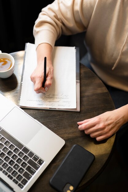 Donna anziana che beve caffè in un bar mentre lavora al suo laptop e scrive sul suo taccuino