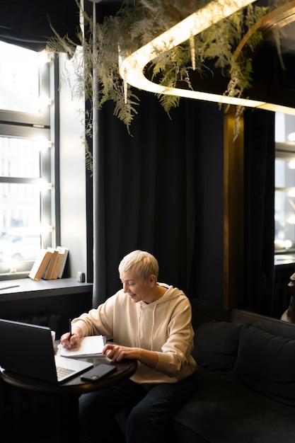 Donna anziana che beve caffè in un bar mentre lavora al suo laptop e scrive sul suo taccuino