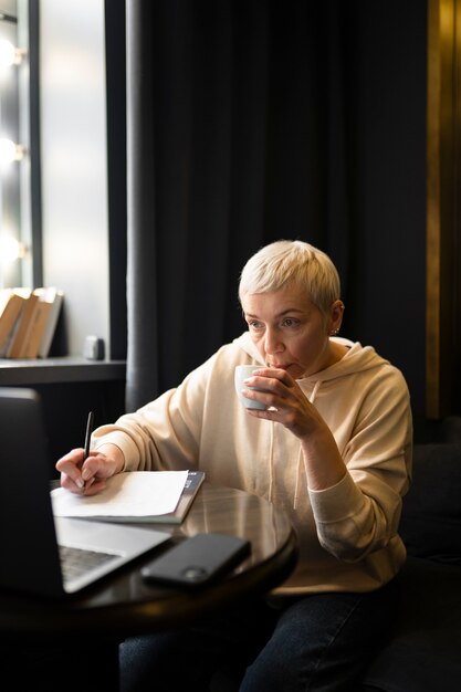 Donna anziana che beve caffè in un bar mentre lavora al suo laptop e scrive sul suo taccuino