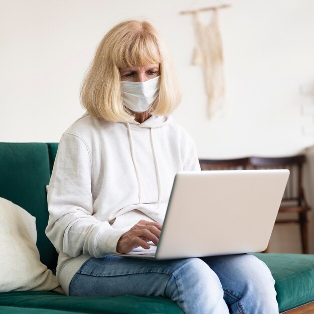 Donna anziana che lavora al computer portatile a casa mentre indossa la mascherina medica