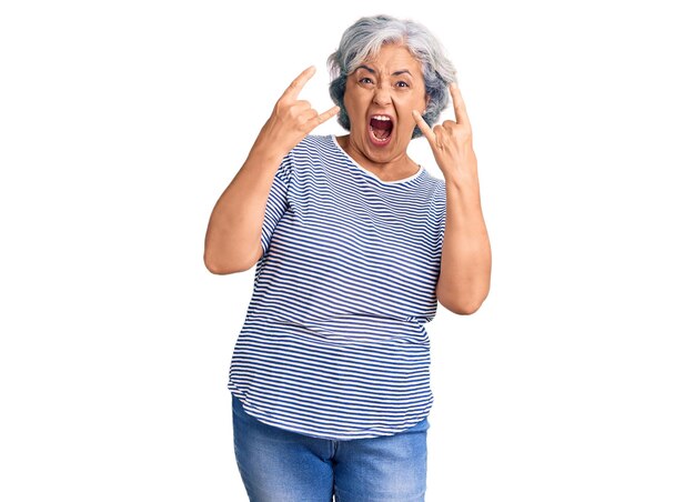 Donna anziana con i capelli grigi che indossa abiti casual a righe che gridano con un'espressione pazza facendo il simbolo della roccia con le mani in alto. stella della musica. concetto pesante.