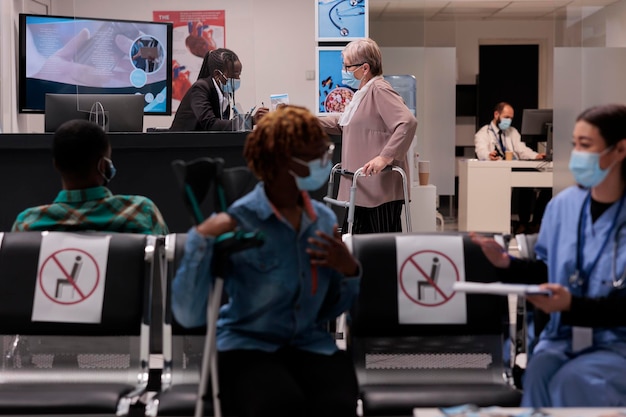 Donna anziana con telaio a piedi che parla con l'addetto alla reception presso la reception dell'ospedale, in attesa di partecipare all'appuntamento presso la struttura medica. Paziente con disabilità fisica e maschera facciale in sala d'attesa.