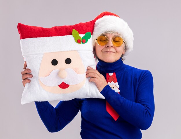 Donna anziana contenta in occhiali da sole con cappello da Babbo Natale e cravatta di Babbo Natale che tiene cuscino di Babbo Natale isolato sul muro bianco con spazio di copia