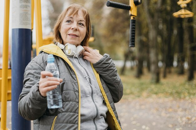 Donna anziana di smiley con le cuffie che bevono acqua dopo aver lavorato all'aperto