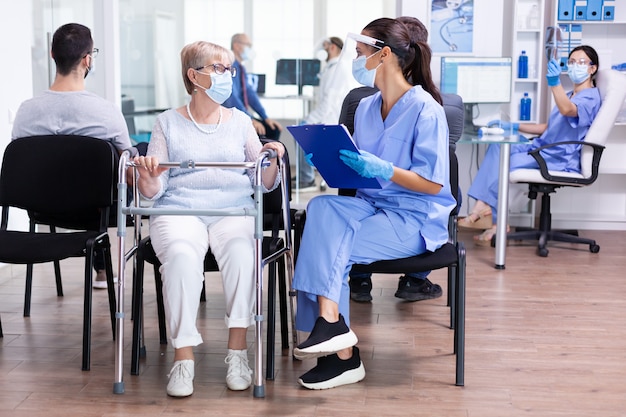 Donna anziana disabile con deambulatore nella sala d'attesa dell'ospedale utilizzando un deambulatore che parla con il personale medico del trattamento per la malattia durante la pandemia di coronavirus