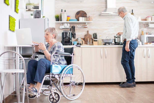 Donna anziana disabile in sedia a rotelle che lavora al computer portatile in cucina. Persona anziana anziana handicappata paralizzata che utilizza la tecnologia web di Internet online moderna di comunicazione.