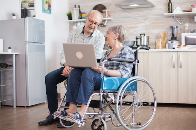 Donna anziana disabile in sedia a rotelle e suo marito che hanno una videoconferenza su tablet pc in cucina. Anziana paralizzata e suo marito durante una conferenza online.