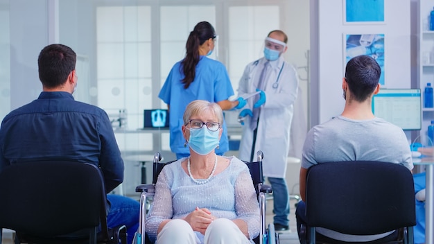 Donna anziana disabile preoccupata seduta in sedia a rotelle nell'area di attesa dell'ospedale per l'esame medico. Vecchia donna che indossa una maschera facciale contro l'infezione da coronavirus.