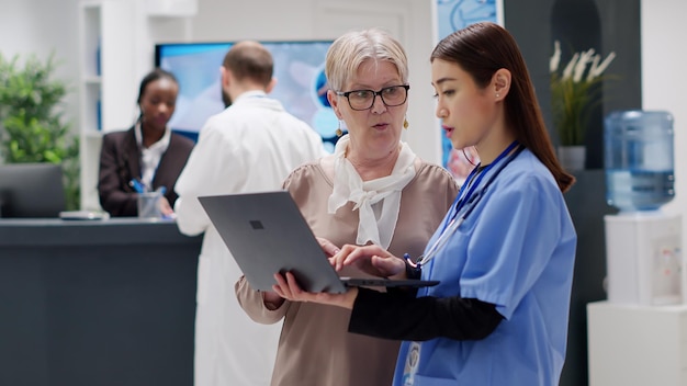 Donna anziana e assistente medico che analizzano il rapporto di diagnosi sul laptop, facendo consulenza alla reception dell'ospedale. Infermiera e paziente che discutono del trattamento e del supporto sanitario. Colpo a mano libera.