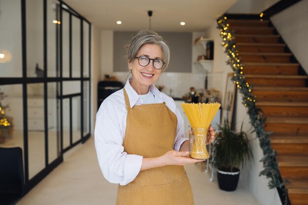 Donna anziana felice che mostra la pasta sulla macchina fotografica Ritratto di signora anziana sorridente che tiene gli spaghetti in piedi nella cucina moderna