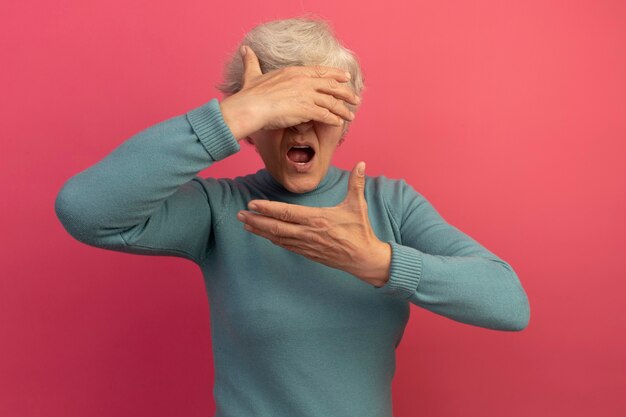 Donna anziana impressionata che indossa un maglione blu a collo alto che copre gli occhi con la mano tenendo un'altra mano vicino al viso isolato sul muro rosa