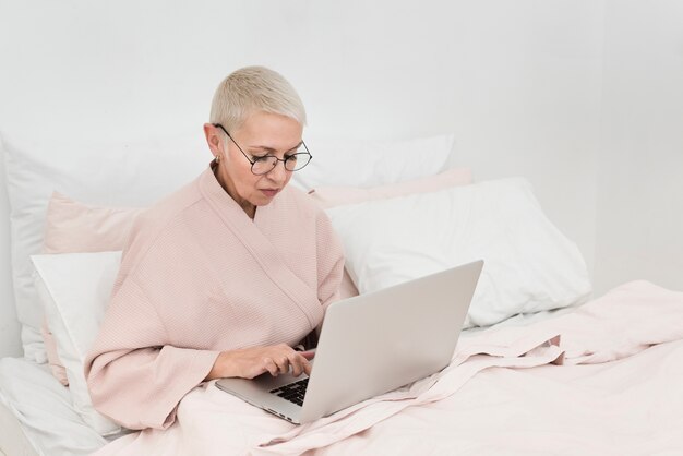 Donna anziana in accappatoio che lavora al computer portatile a letto