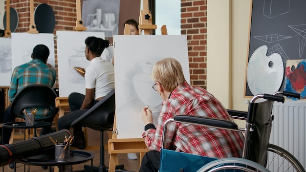 Donna anziana in sedia a rotelle che disegna uno schizzo di vaso con matita su tela e cavalletto, frequentando il programma di lezione d'arte con studenti e insegnante. Persona con disabilità che impara a disegnare per crescere.