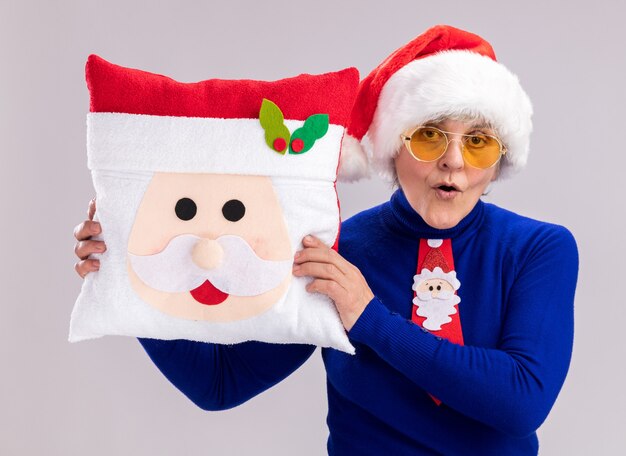 Donna anziana sorpresa in occhiali da sole con cappello babbo natale e cravatta babbo natale che tiene cuscino babbo natale isolato su muro bianco con spazio copia