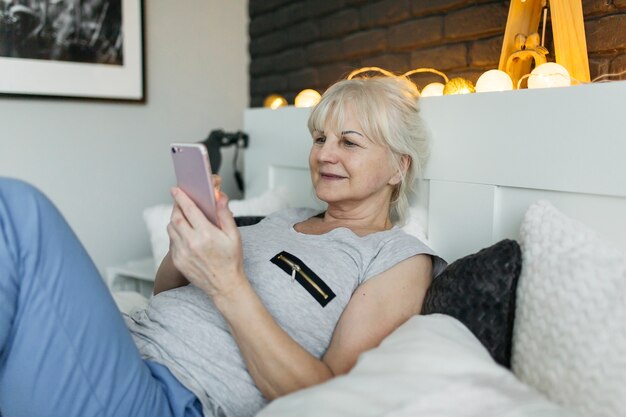 Donna anziana sorridente che per mezzo dello smartphone sul letto