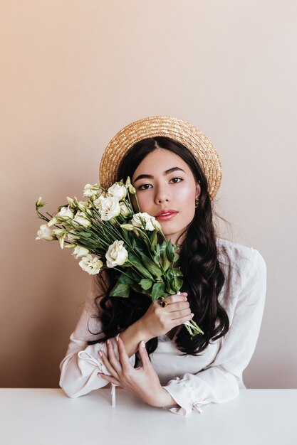 Donna asiatica alla moda in cappello di paglia che tiene eustomas bianchi. Vista frontale della bella donna riccia con bouquet di fiori su sfondo beige.