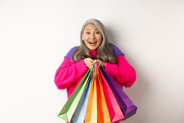 Donna asiatica anziana alla moda shopaholic, abbracciando le borse della spesa e sorridendo gioiosa, acquistando con sconti, in piedi su sfondo bianco