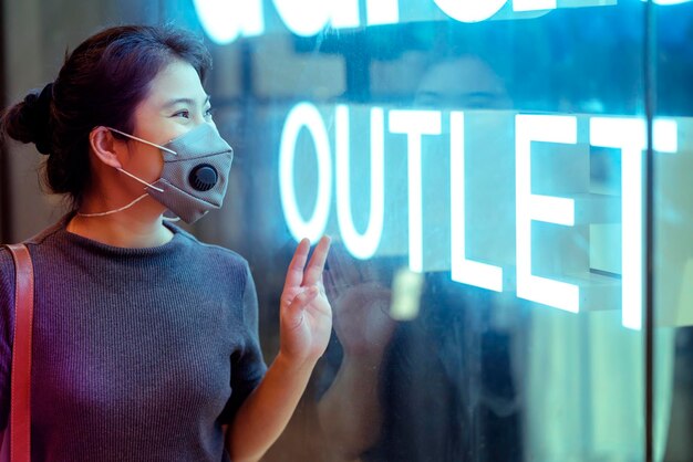 Donna asiatica attraente allegra che indossa una maschera protettiva guardando la vetrina della vetrina della vetrina della segnaletica di vendita dell'outlet nuovo concetto di stile di vita normale