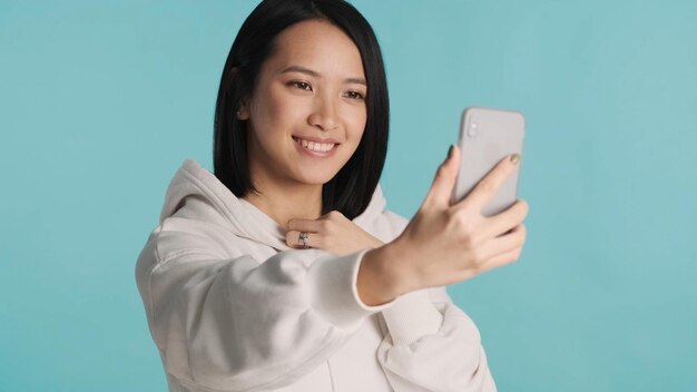 Donna asiatica che sembra carina sinceramente sorridente prendendo selfie sullo smartphone su sfondo blu