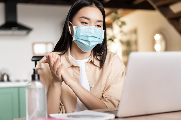 Donna asiatica che si lava le mani con un disinfettante per le mani che indossa una maschera medica per il viso e lavora al video del laptop ...