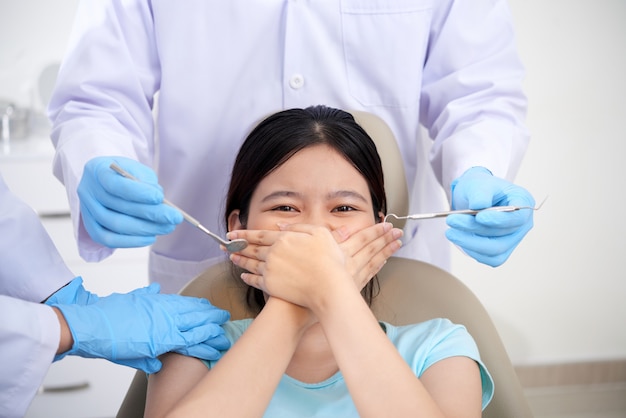 Donna asiatica che si siede nella clinica dentale, coprendo la bocca con entrambe le mani e medici in piedi accanto