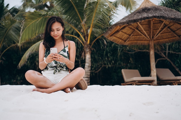 Donna asiatica che si siede sulla spiaggia e che per mezzo del telefono