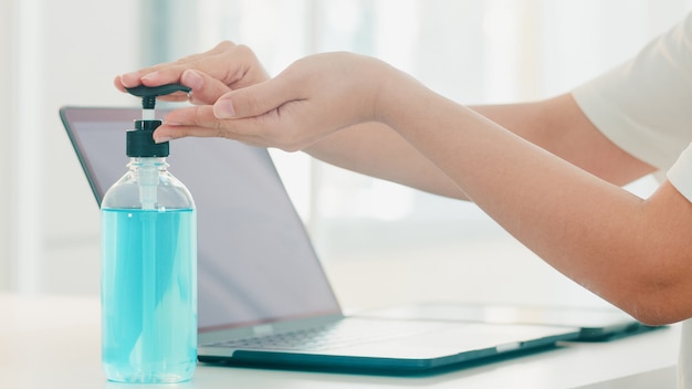 Donna asiatica che usando la mano del lavaggio del disinfettante della mano del gel dell'alcool prima del computer portatile aperto per proteggere il coronavirus. Le donne spingono l'alcool per pulire per l'igiene quando il distanziamento sociale rimane a casa e il tempo di auto-quarantena