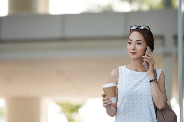 Donna asiatica con caffè da asporto che cammina in strada e che parla sul telefono