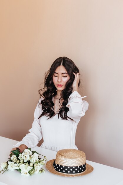 Donna asiatica con fiori bianchi che toccano i capelli ricci. Studio shot di gioiosa signora giapponese seduta a tavola con bouquet di eustoma.