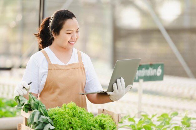 Donna asiatica dell'agricoltore che lavora con il computer portatile nella fattoria idroponica di verdure organiche. Proprietario dell'orto di insalata idroponica che controlla la qualità della verdura nella piantagione di serre.