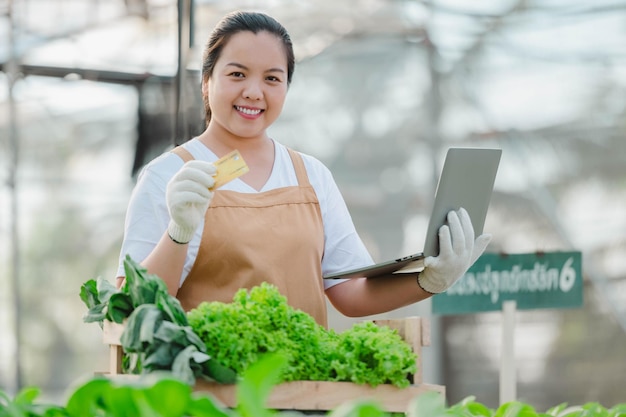 Donna asiatica dell'agricoltore che lavora con il computer portatile nella fattoria idroponica di verdure organiche. Proprietario dell'orto di insalata idroponica che controlla la qualità della verdura nella piantagione di serre.