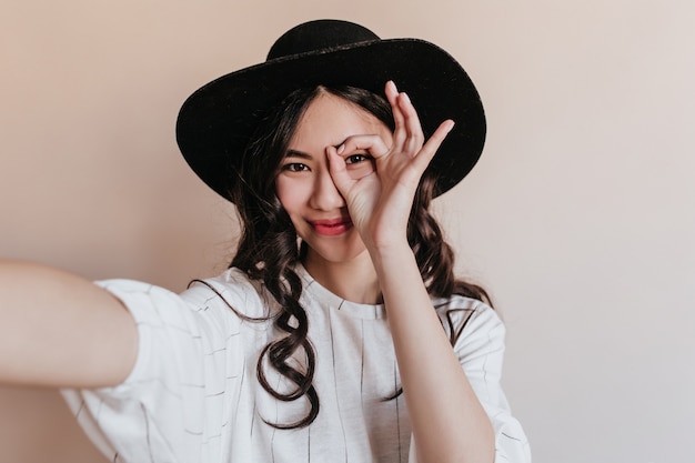 Donna asiatica divertente che posa con il segno giusto. Modello giapponese in cappello prendendo selfie su sfondo beige.